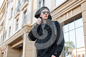 Trendy young hipster man in a stylish hooded sweatshirt in dark sunglasses in a cap with a backpack is standing in a city