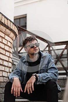 Trendy young man hipster in stylish denim blue clothes trendy in a black sunglasses sits on a vintage staircase