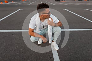 Trendy young man in vintage blue jeans in a fashionable T-shirt in white stylish sneakers with a hairstyle relaxes on the asphalt