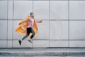 Trendy young man in yellow raincoat jumping in the air on wall street urban background