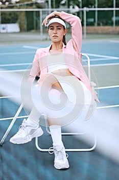 Trendy young tennis player wearing a pink jacket while sitting on chair on a tennis court. Young hispanic sportswoman