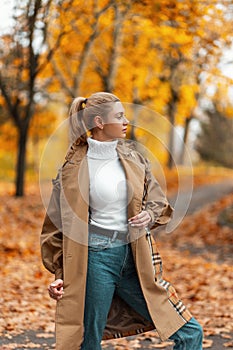 Trendy young woman blonde in an elegant coat in a vintage sweater in trendy jeans stands in the park and enjoys the autumn