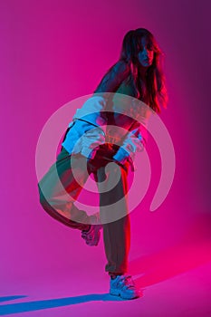 Trendy young woman dancer in a youth fashion tracksuit in stylish sneakers dancing hip-hop in the studio with bright pink neon