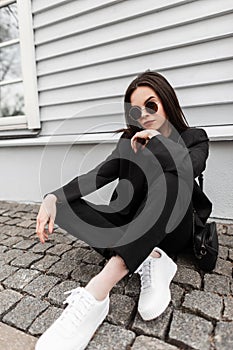 Trendy young woman hipster in sunglasses in fashionable youth black clothes in white trendy sneakers with leather handbag sit on