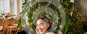 Trendy young woman sitting in cafe and smiling at camera, drinking coffee and using laptop, working remotely, studying