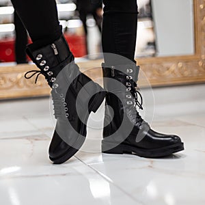 Trendy young woman stands on a tile in store in black cool leather lace-up boots with vintage jeans. Close up of female legs in