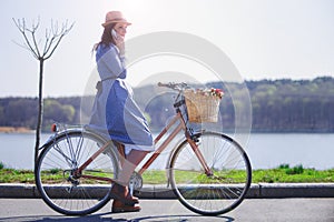 Trendy young woman stop to riding on her vintage bike with basket of flowers while focused chatting or talk on smart phone outside