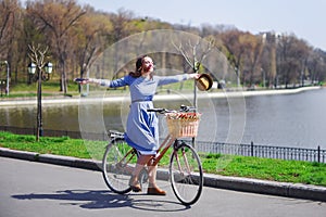 Trendy young woman stop to riding on her vintage bike with basket of flowers while focused chatting or talk on smart phone outside