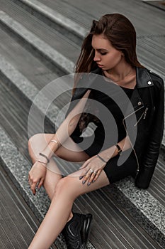 Trendy young woman in a stylish dress in a fashionable leather jacket in autumn black boots relaxes sitting on a stone staircase