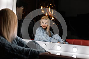 Trendy young woman in stylish knitted clothes sits and looking in a vintage white mirror in a room with Christmas decorations.