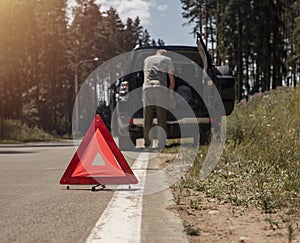 Triangle caution sign on road near broken car with driver on background