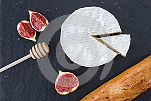 Triangular piece of camembert cheese and a formed cheese, figs in honey and a wooden spoon for honey and browned baguette on a gra