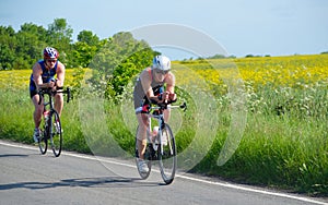 Triathletes on road cycling stage of triathlon fields and trees in background.
