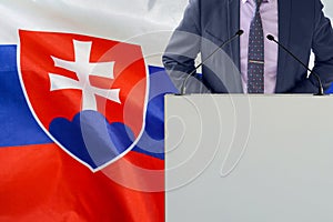 Politická osobnosť stojaca na pódiu konferencie na pozadí vlajky Slovenska. Muž hovorca na Slovensku