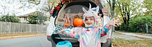 Trick or trunk. Happy baby in unicorn costume celebrating Halloween in trunk of car. Cute toddler preparing for October holiday