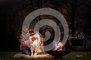 Tricolor Australian Shepherd sits next to a young woman by the campfire. In winter, snow on the grass. The fire is