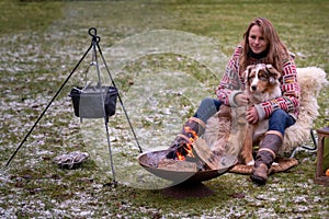 Tricolor Australian Shepherd sits next to a young woman by the campfire. In winter, snow on the grass. The fire is
