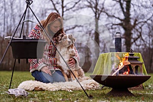 Tricolor Australian Shepherd sits next to a young woman by the campfire. In winter, snow on the grass. The fire is burning.