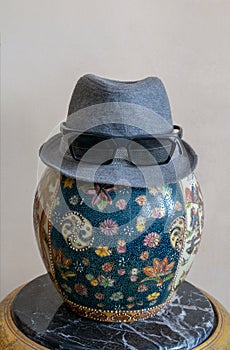 Trilby hat wearing sunglasses over antique decorated Chinese ceramic jar vase over antique black marble table