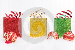 Trio of glittery gift bags in a row on a white background and fur with red and gold Christmas ornaments