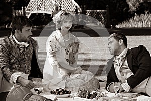 Trio of people dressed in vintage clothing on picnic blanket. Gentlemen stare off as beautiful lady looks to camera