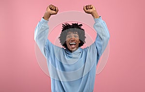 Triumphant black teen guy lifting arms above his head, shouting in excitement on pink studio background