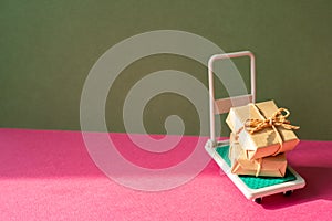 Trolley cart with gift boxes on pink and khaki green background. Parcel Delivery Concept