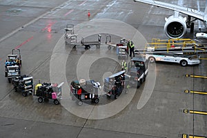 Trolleys full of luggage are standing by the airplane and two airport workers are loading them by conveyor onto the plane.