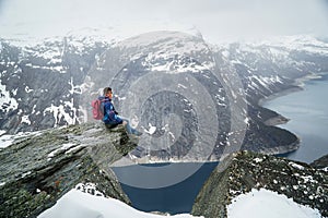 Trolltunga cliff under snow in Norway. Scenic Landscape. Man traveller sitting on edge of rock and looking at distance