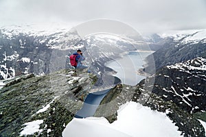 Trolltunga cliff under snow in Norway. Scenic Landscape. Man traveller sitting on edge of rock and making photos of