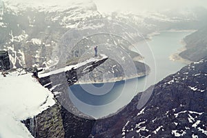Trolltunga cliff under snow in Norway. Scenic Landscape. Man traveller standing on edge of rock and looking down. Travel