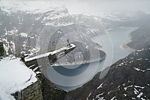 Trolltunga cliff under snow in Norway. Scenic Landscape. Man traveller standing on edge of rock and looking down. Travel