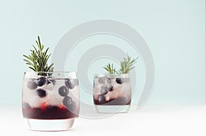 Tropical alcoholic shots of sweet berry liquor with ice cubes, blueberry, rosemary on pastel soft light green background.