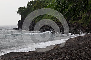 Tropical beach on storm - cape with black rocks, lush green forest and   grey waves of sea with foam in rain.  Trip in rain season