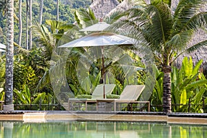 Tropical beach with swimming pool, coconuts palm trees, two deck chairs and umbrella near sea, Thailand