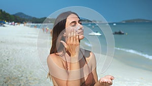 On tropical beach woman applying sunscreen to face protecting skin from sun's rays. Strokes of sunscreen prioritizes