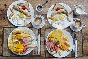 Tropical breakfast of fruit, coffee and scrambled eggs and banana pancake for two on the beach near sea. Top view, table setting
