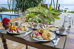 Tropical breakfast of fruit, coffee and scrambled eggs and banana pancake for two on the beach near sea. Top view, table setting