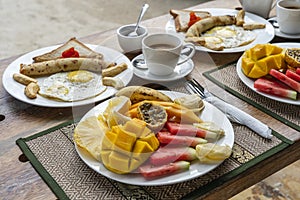 Tropical breakfast of fruit, coffee and scrambled eggs and banana pancake for two on the beach near sea. Top view, table setting