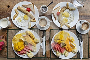 Tropical breakfast of fruit, coffee and scrambled eggs and banana pancake for two on the beach near sea. Top view, table setting