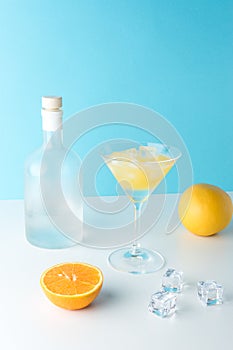 Tropical cocktail in martini glass with ice cubes and bottle of alcohol on white table. Minimal summer drink composition