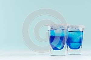 Tropical fresh alcohol cocktail with blue curacao liqueur, ice cube, sugar rim in two misted shot glass on soft light white.