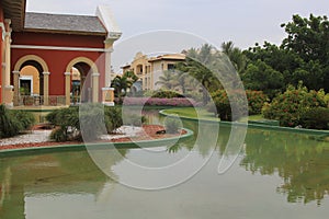 Tropical garden with green grass, colorful houses in the background, a small lake with calm water and reflections Abrir no Google photo