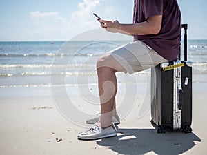 Tropical sea, Young man sitting on Travel suitcase and using smart phone at the beach. Summer holiday traveling concept design