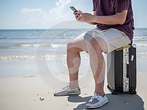 Tropical sea, Young man sitting on traveling bag and using smart phone at the beach. Summer holiday traveling concept design