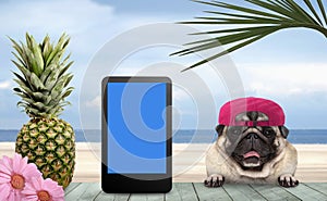 Tropical summer pug dog with cap and tablet, with paws on vintage green wooden table and sea and beach on background