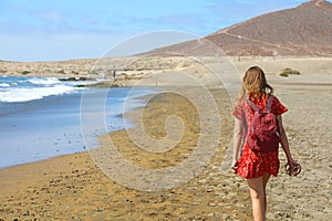 Tropical traveling. Young woman with red dress and backpack walking barefoot by sea beach enjoying landscape