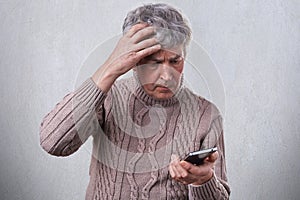 A troubled mature man holding his hand on his gray head while using smartphone reading some news in the Internet. A man having sho