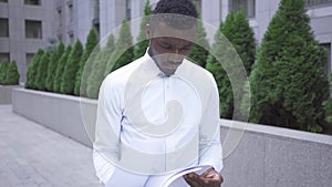 Troubled young African American man in white shirt looking through papers standing on the city street. Male businessman