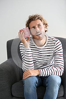 Troubled young man with piggy bank sitting in armchair at home